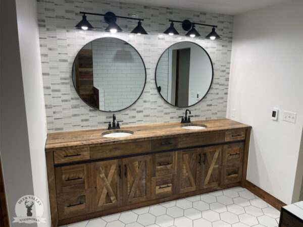 Rustic barnwood vanity with double sinks set in a large barnwood top, three drawers at each end and in the center, a cabinet behind barnwood doors with recessed center panels below each sink, and antiqued blackened metal strap on the face and outside corner brackets on the countertop.