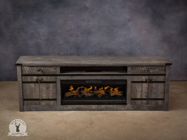 Rustic grey stained reclaimed barnwood media cabinet with center electric fireplace, large solid barnwood top, center shelf above fireplace, storage drawer and double cabinet doors with railroad spike knobs at each end of the cabinet and antiqued blackened metal accent straps with integral steel rivets.