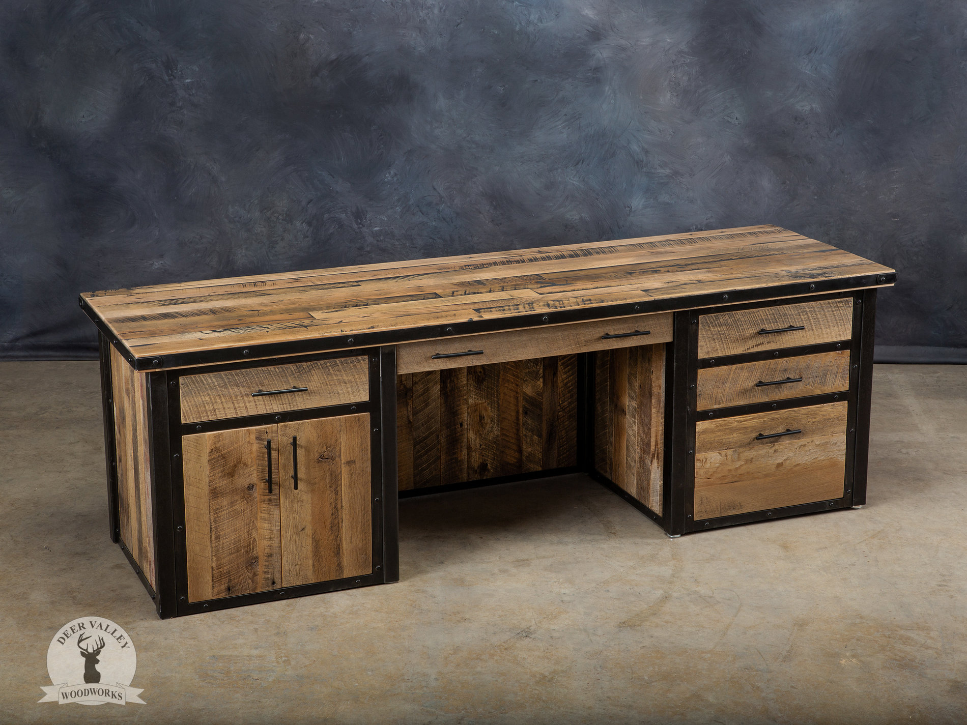 Industrial executive barnwood office desk has a large desktop rapped in an antiqued steel strapping and secured with welded steel rivets, privacy panels, a center drawer, three storage drawers, a file drawer and double door storage cabinet all surrounded by a blackened welded metal frame.